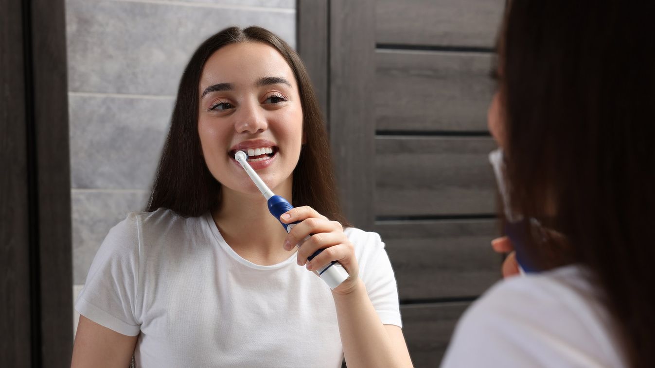 Young,Woman,Brushing,Her,Teeth,With,Electric,Toothbrush,Near,Mirror